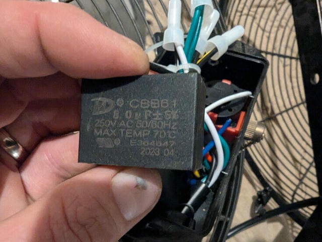 Fan capacitor The author holding a CBB61 capacitor, with some wires, a pull chain switch, and some fan housing elements visible in the background